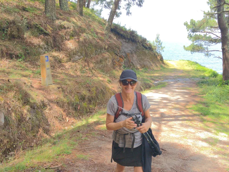 A woman walks along a path by the sea, holding a camera and wearing a backpack, with trees in the background.