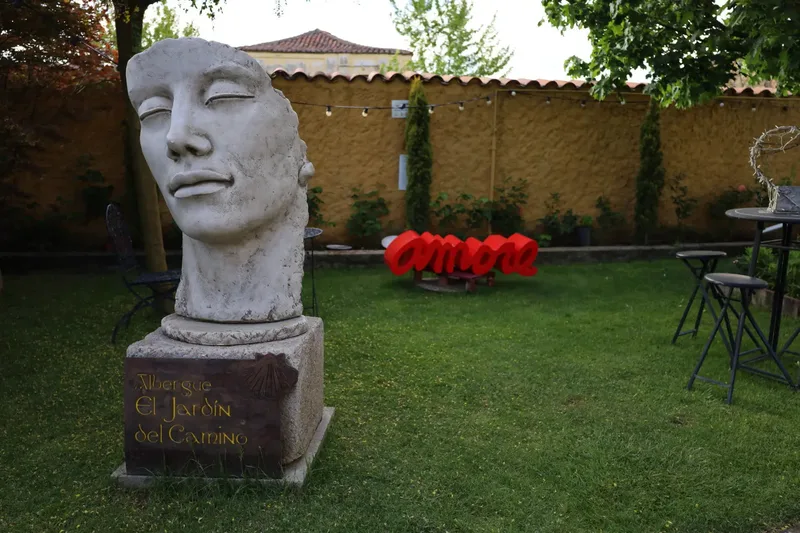 A large stone head sculpture stands in a garden with green grass, a red "amor" sign, and black chairs nearby.