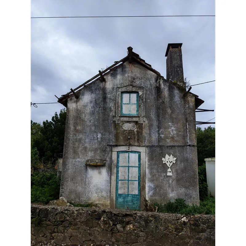 A weathered two-story house with blue windows, a slanted roof, and a decorative white symbol on the wall.