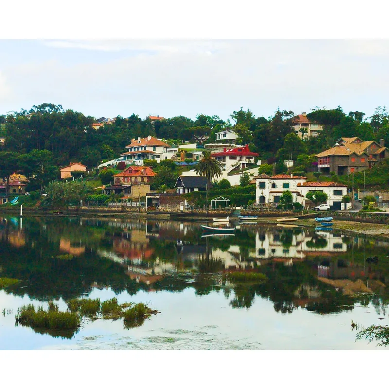 Houses line a hillside next to a calm water body, reflecting buildings and greenery. Boating equipment is visible.