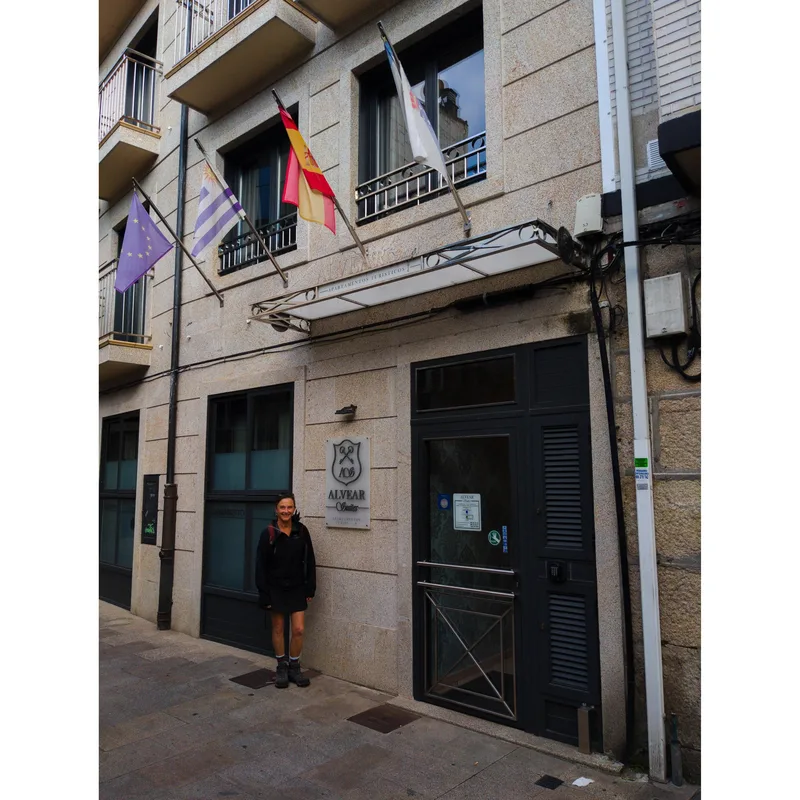 A woman stands by the entrance of a building with flags and a sign reading "ALVEAR.
