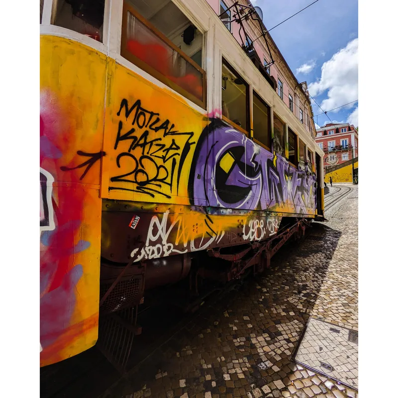 A colorful tram covered in graffiti parked on a cobblestone street with buildings in the background and blue sky.