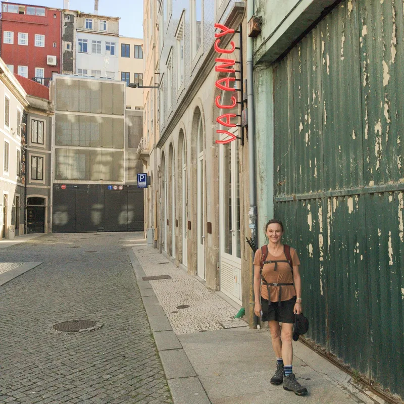 A woman stands in a cobblestone street with a "VACANCY" sign, alongside weathered buildings and a parking sign.