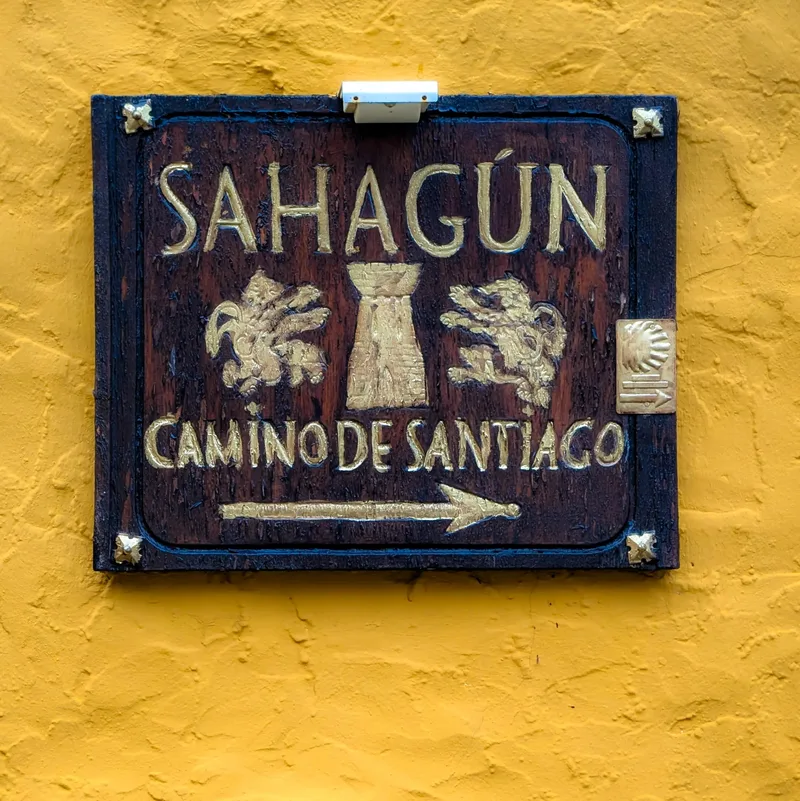 Wooden sign with gold lettering reading "Sahagún Camino de Santiago" on a yellow wall, with directional arrow.
