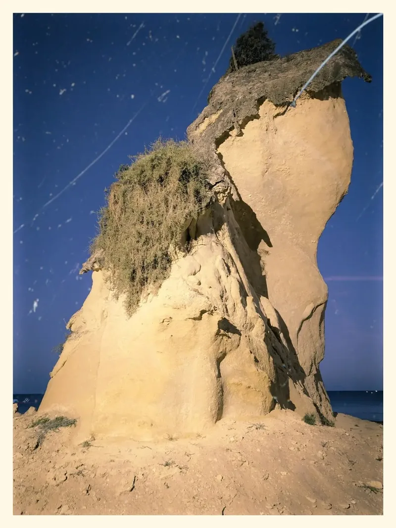 A tall, sandy rock formation with vegetation on top, set against a blue sky and ocean.