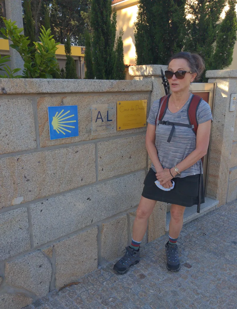 A woman in hiking attire stands by a stone wall with signs for "Casa da Pria" and a yellow sun symbol.