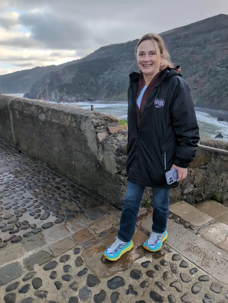 A smiling woman stands on a stone pathway near a cliff overlooking an ocean landscape, wearing colorful sneakers.