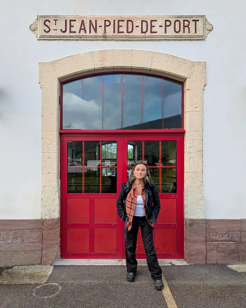 A woman stands in front of a red door with a sign reading "S. JEAN-PIED-DE-PORT." in a rural setting.