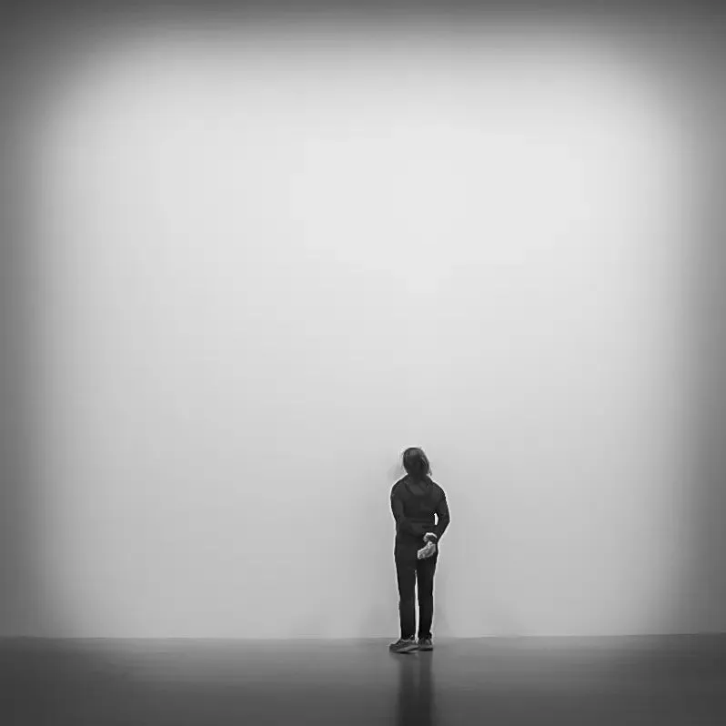 A person stands with their back to the viewer, facing a large, plain white wall.