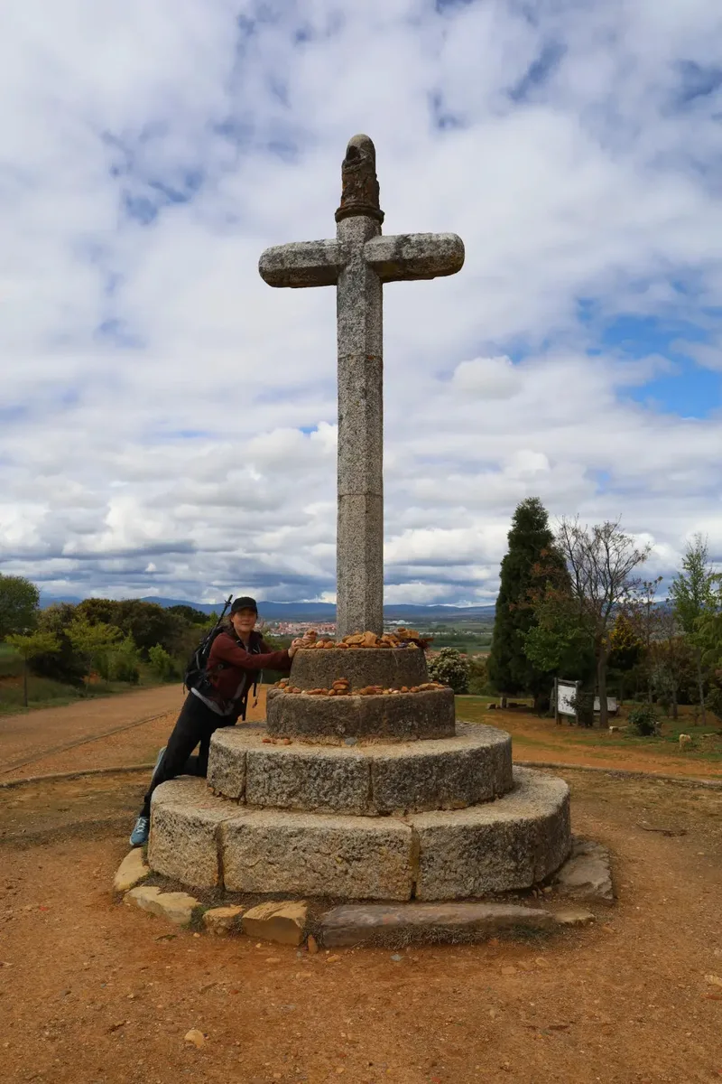 A person embraces a stone cross on a hill, with clouds and trees in the background.