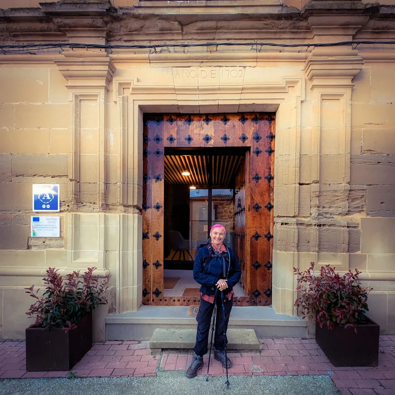 Person with a walking stick stands in front of a large wooden door featuring black decorative hinges.