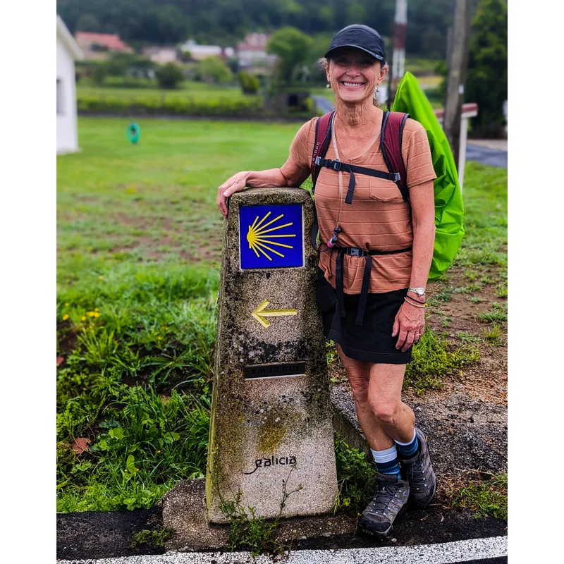 A woman stands beside a stone marker with a shell symbol, indicating a Camino de Santiago route in Galicia.