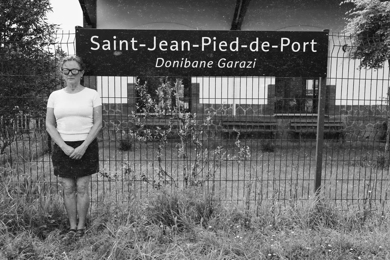 A woman stands beside a sign reading "Saint-Jean-Pied-de-Port Donibane Garazi," with a building and grass nearby.