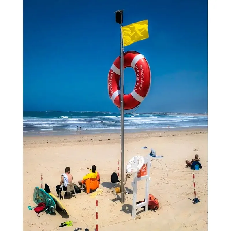 Lifebuoy on a pole with a yellow flag, beachgoers sitting on sand, surfboards nearby, and ocean waves in background.
