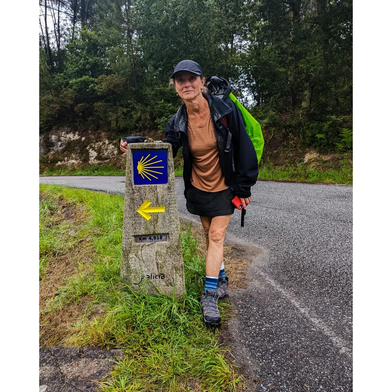 A woman stands by a stone milestone with a yellow arrow indicating 4.918 km along a winding road in a forested area.